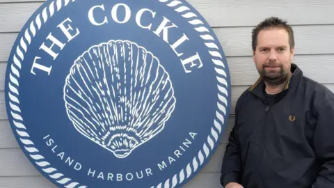 A man stood next to a sign saying 'The Cockle Island Harbour Marina'