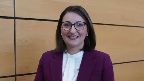 Ema Cubitt standing in front of a wooden panelled wall, smiling at the camera.  She has shoulder-length dark hair and glasses and is wearing a purple suit jacket over a white blouse. 