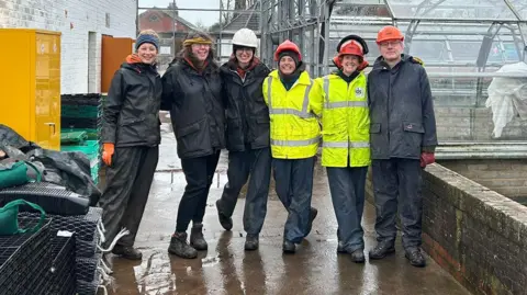 Living Well Five people, some wearing hard hats and hi-vis jackets stand in front of a greenhouse. It is raining and they are all wearing waterproof clothing 