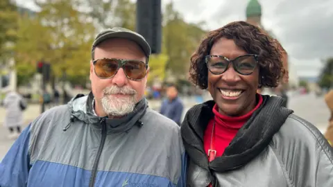 Andy and Alisha Lewis smiling successful  beforehand   of Belfast City Hall. Andy is wearing a raincoat, headdress  and sunglasses portion    Alisha is wearing glasses, a reddish  jumper and achromatic  coat. 