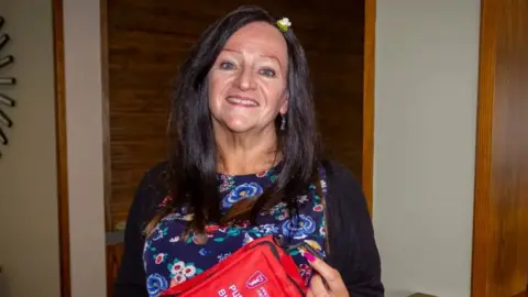 Lynne Baird The image shows Dr Lynne Baird standing indoors, holding a bright red first-aid pouch with “Public Bleed Control Kit” printed on the front. She is wearing a dark cardigan over a patterned top with floral designs. The background features wooden wall panels and a doorway.