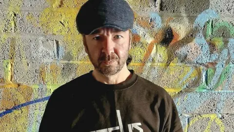 Facebook Bobby McNamara - a man wearing a flat cap, with a beard, wearing a dark top and standing in front of a wall with graffiti sprayed on it