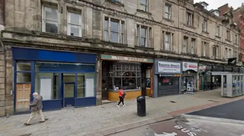 Google The Posada, featuring a partly brown and green sign, is one of the premises visible as being on the ground floor on a road. Two people are walking on the pavement in front and part of the road is also on the photo.