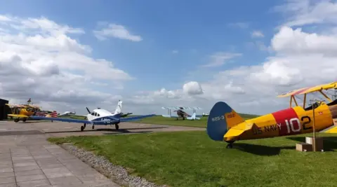 A blue, red and yellow plane and a white and blue plane stationary at Compton Abbas Airfield. It's a sunny day and the sky is blue