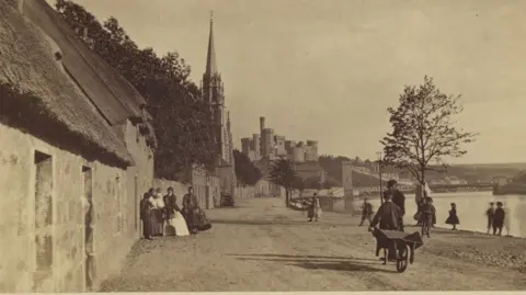 Getty Images The sepia image shows an Inverness Riverside scene. On the left are cottages with thatched roofs. A group of women in long dresses are gathered near one of the cottages. They are standing on a rough, dusty road. A number of other people are on the road, including a boy who is pulling a wooden wheelbarrow and is talking to a man with a bushy beard and is wearing a flat cap. The River Ness flows on the right of the image and in the distance is a bridge and Inverness Castle.