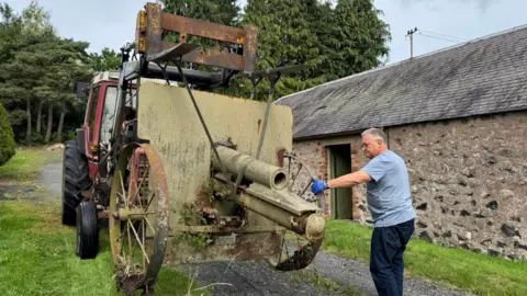 Great War Huts. A rusty short-barrelled 10.5cm howitzer gun with its barrel facing the viewer. It has been raised up by a red tractor. Kev Smith is holding its wheel on the right. Beyond it is a low stone-built barn. 