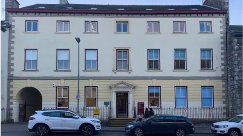 Strickland House is a Georgian building which has large windows and decorative panels. It is painted cream, white and maroon and looks well-cared for. There are two cars parked outside. 