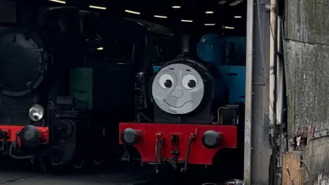 A close-up of Thomas a locomotive at Nene Valley Railway. The blue locomotive has a smiling face painted on the front of its engine. It is sitting in a shed in sidings. 