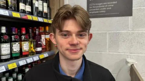 A head and shoulders view of shop assistant Reagan Bennie standing next to shelves full of spirits. He has brown hair in a curtains-style cut, and wears a blue shirt and black fleece.