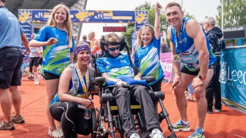 PA Media Rob Burrow, in a wheelchair, alongside wife Lindsey, daughters Macy and Maya and Kevin Sinfield on a red track at Leeds.