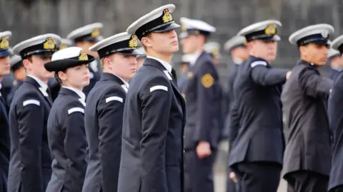 A cohort of Royal Navy recruits stands on parade during a passing out ceremony. They are all wearing uniform with hats on.