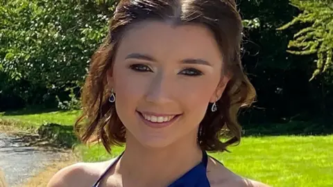 Family photo Jessica Gallagher smiling at the camera on a sunny day. She has wavy brown hair which falls to just above her shoulders. She is wearing a blue formal dress and standing in a lawned area with trees in the background.