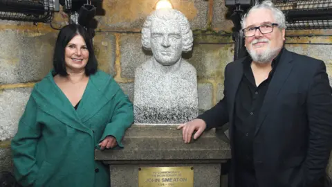 Wayne Perry Miranda Housden (left) and Steve Conway (right) are pictured in front of the plaque. Miranda is wearing a long green jacket. She has short black hair. Steve is wearing a black shirt with a black blazer on top of it. He has grey hair and a grey beard with black glasses.