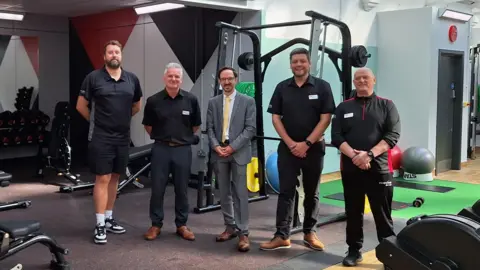 Five men stand in front of a selection of gym equipment.