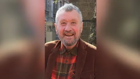 A man with grey hair and a grey beard smiling at the camera. He is wearing a brown jacket and a red and black checked shirt