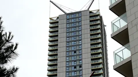 A close-up, cropped shot of the upper floors and distinctive V-shaped crown of a modern grey residential skyscraper, flanked by a pine tree on the left and the glass balconies of a neighbouring building on the right.