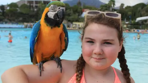 Charly Bisset standing next to a swimming pool with a parrot on her right arm. Her hair is in braids and she has sunglasses on top of her head.