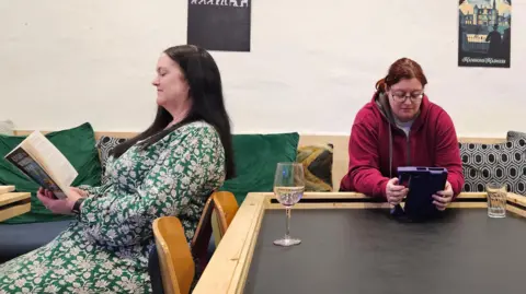 Two women, sat facing away from each other, both with dark hair. One is reading a paperback book, the other a purple tablet.