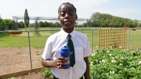 Qays Najm/BBC A teenage boy stands in a school field and smiles at the camera. He is holding a blue bottle. He is wearing a white shirt and green tie. 
