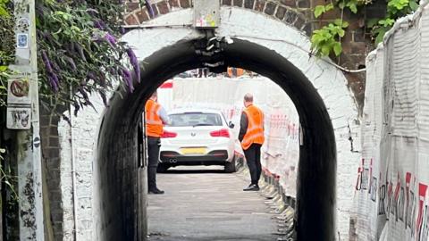A narrow walled footpath passing under a bridge, with a white BMW car stopped in the middle and two men in orange high vis jackets looking at it