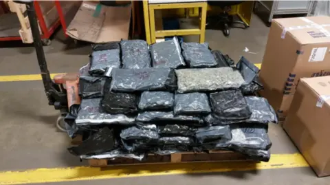 Image shows a stack of vacuum-packed blocks of cannabis on a trolley in an airport warehouse