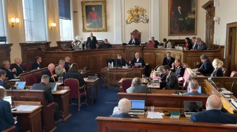 BBC A group of men and women in suits sitting in a court setting. 