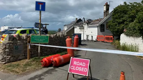 The entrance to the Worm's Head Hotel. There is a road closed sign just at the entrance with police table above it. There is orange canister lying on the ground. The roof of the hotel has been damaged by the fire. There is a police car parked in front of the hotel