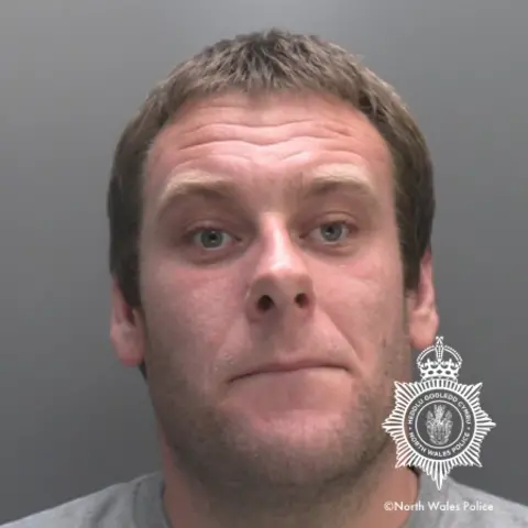 North Wales Police Custody head shot of Michael Owen Williams, a white male man with a heavily wrinkled forehead, light short brown hair, and stubble. He is wearing a grey top, and there is a large North Wales Police badge emblem and copyright watermark on the bottom righthand of the image