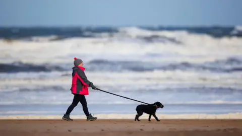 A man wearing a red coat, hat, black trousers and walking boots, walks along a sandy beach. He is walking a black dog. He is using one hand to hold the lead which is stretched out in front of him. Waves are crashing in the background.