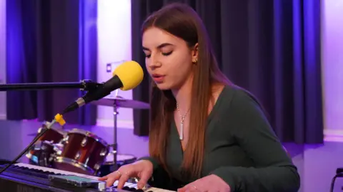 Gordano School A teenage girl playing a piano while singing into a yellow microphone. She has long, straight brown hair and wears a dark green long-sleeved top with a silver pendant necklace. A red drum kit and some navy-blue curtains can be seen in the foreground.