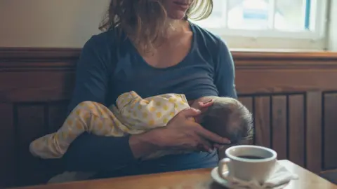 In a cafe with dark wooden backed benches, a woman in a blue top breastfeeds a baby in a yellow and white babygrow. A cup of coffee in a white cup sits on a  table in front of them.