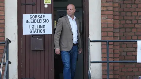 Dan Thomas stepping out of a door in jeans, a jacket and a shirt, with a sign that reads polling station next to him.