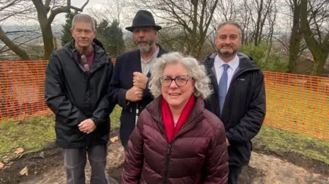 A woman is stood in front of three men. There is an orange plastic mesh fence behind them and trees in the distance. The woman has grey, white hair nearly to her shoulders and wears a purple coat and red scarf. Two of the men have beards and all wear coats.