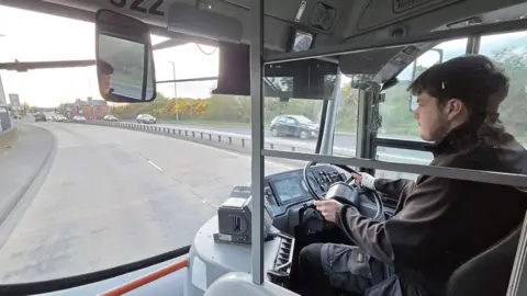 Luke Wilson Luke Wilson driving a bus. He has brown hair, wearing a black jacket and black trousers. He is driving along a dual carriageway with cars on the other side.