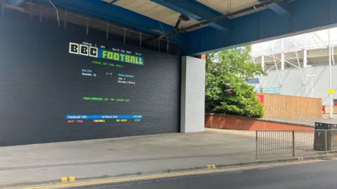 Middlesbrough Council Mural in the style of the former television teletext service Ceefax. Set against a dark grey brick wall it states in red, yellow, green, blue and white letters: BBC Football: League Cup Final Result. Bolton 1 -2 Middlesbrough. Middlesbrough lift the league cup