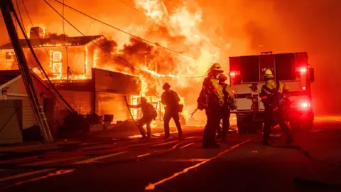 Kyle Grillot/Bloomberg via Getty Images Siluet petugas pemadam kebakaran berkumpul di depan mobil pemadam kebakaran di samping rumah yang terbakar di kawasan Pacific Palisades