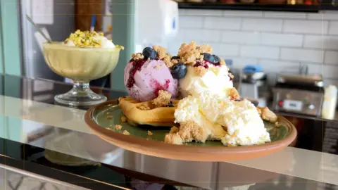 Sobre una encimera hay dos copas de helado. Una tiene helado de vainilla y frutos rojos, con arándanos y una cobertura crujiente. La otra es de pistacho verde con café expreso.