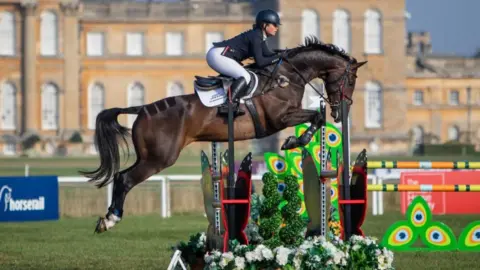 Saffron Cresswell A female jockey riding a dark brown horse as it jumps over fences, with a large stately building behind. 