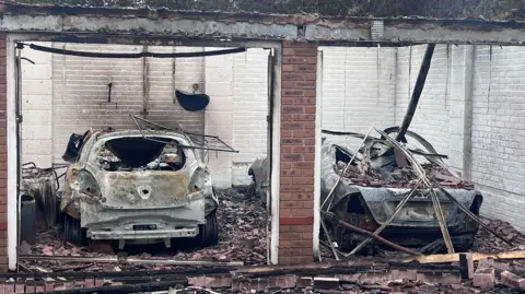 Two cars and a garage which have been completely destroyed by fire. The cars are grey and caved in and there is much material around them.