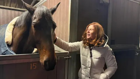 Una mujer acaricia a un caballo marrón en la cabeza. Tiene el pelo rojo y el pelaje blanco.
