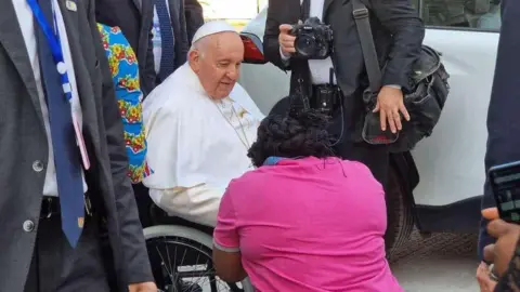 Kitsita Ndongo Rachel Pope Francis wearing white vestments is seated on a wheelchair, looking at Kitsita Ndongo Rachel who is wearing a pink top and is kneeling down. There are people standing around them wearing grey suits and one is carrying a black camera bag  and is holding a black Nikon camera.