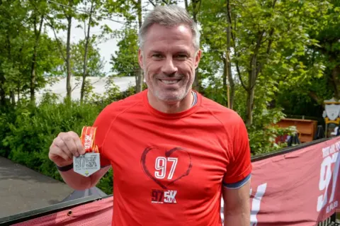 Former Liverpool footballer Jamie Carragher wearing a red BTR Run For The 97 sports top on smiles as he holds a red and silver 97 5K medal up on a sunny day. 