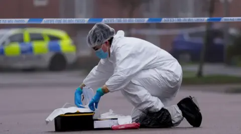 PA Media A forensics officer in a white suit, blue gloves, a facemask and a hairnet kneels on the road in front of police tape. There is a case full of equipment in front of them.