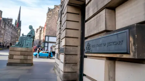 A general view of the outside of the High Court in Edinburgh. It is a stone building and the view is looking up the street to a statue and other buildings