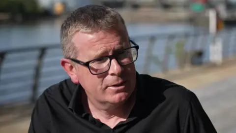 BBC Dr John Mulligan wearing glasses and a black shirt sitting on a bench with railings and water in the background