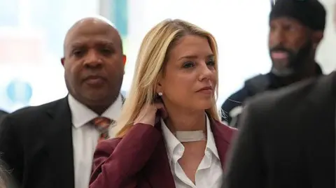 A woman with long blonde hair, flanked by men in suits, adjusts the collar of her white shirt under a maroon jacket. She has a bandage at the base of her throat