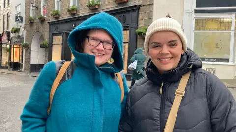 Rachel Gilbert and Martine Ellis are standing in a street and smiling at the camera. Rachel is on the left and is wearing a turquoise coat. We can see the brown straps of her rucksack. On the right is Martine and she is wearing a white woolly hat, and a dark puffy coat with a zip.