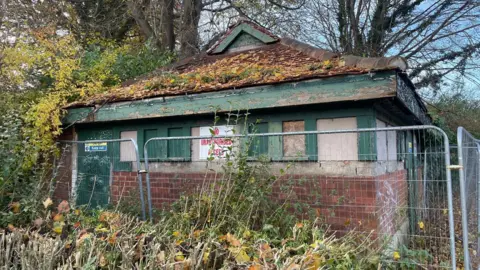Durham County Council The old toilet block is a one story brown-brick building with a triangle roof and green door and window panes. There is a pile of yellow tree leaves on the roof. NO UNAUTHORISED ACCESS is written in red on a sign attached to the building. A grey metal fence is surrounding the block.
