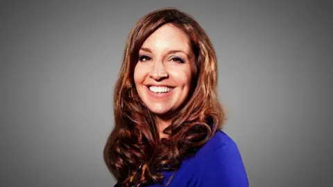 A woman with long brown hair smiles at the camera. she is wearing a bright blue top and there is a grey background.