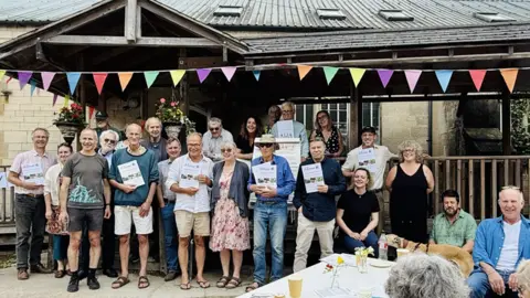 Stroud Common Wealth A group of middle-aged people standing in front of a large building with a wooden covered walkway. Some of them are holding pamphlets up. There is a table just visible in the foreground with small jars holding tiny flowers